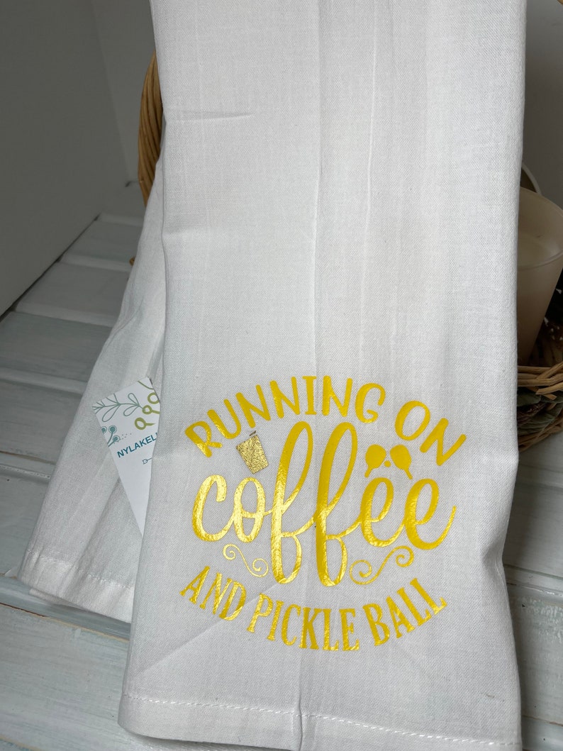 Op de afbeelding: Witte keukendoek met de tekst "Running on coffee and pickle ball" in gouden letters.