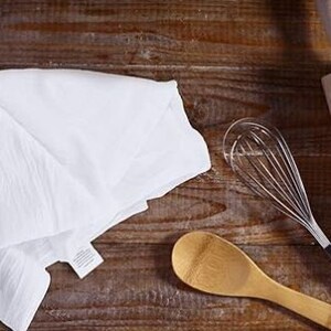 Op de afbeelding: Een witte keukendoek, een houten lepel, een houten deegroller en een zilveren garde op een houten oppervlak.