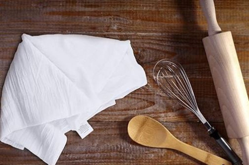 Op de afbeelding: Een witte keukendoek, een houten lepel, een houten deegroller en een zilveren garde op een houten oppervlak.