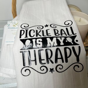 Op de afbeelding: Een witte keukendoek met zwarte tekst die "Pickleball is my therapy" luidt, met een ster en swirls aan beide kanten.