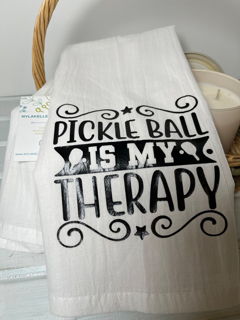Op de afbeelding: Een witte keukendoek met zwarte tekst die "Pickleball is my therapy" luidt, met een ster en swirls aan beide kanten.