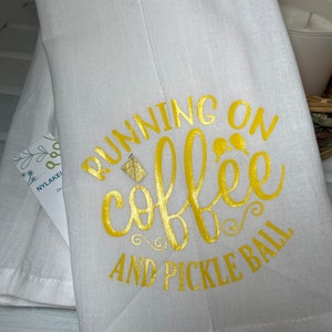Op de afbeelding: Witte keukendoek met gouden opschrift "Running on coffee and pickle ball".
