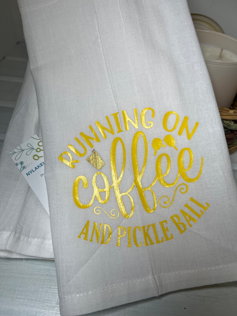 Op de afbeelding: Witte keukendoek met gouden opschrift "Running on coffee and pickle ball".
