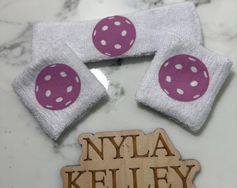 May include: A white terry cloth headband and two matching wristbands, each featuring a purple pickleball graphic with white circular holes. Below the accessories is a light-colored wooden cutout sign with the text NYLA KELLEY engraved in black.