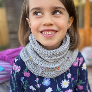 May include: A young girl wearing a gray crocheted cowl with colorful beads. The cowl is folded over twice and has a relaxed fit. The girl is wearing a blue and white floral shirt.