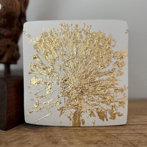Könnte beinhalten: Eine weiße quadratische Fliese mit einem Design aus Blattgold in Form eines Löwenzahns. Der Löwenzahn befindet sich in der Mitte der Fliese und hat eine detaillierte, strukturierte Optik.