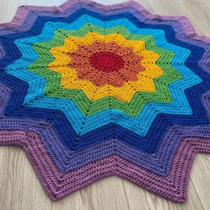 May include: A rainbow crochet star blanket with a purple border. The blanket is made with a variety of colors, including red, orange, yellow, green, blue, and purple.