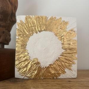 Könnte beinhalten: Eine weiße quadratische Leinwand mit einem goldenen Kreis in der Mitte. Die goldene Farbe ist strukturiert und hat einen erhabenen Effekt.