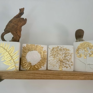 Könnte beinhalten: Vier weiße quadratische Fliesen mit gepressten botanischen Motiven aus Blattgold. Die Fliesen zeigen einen Farn, eine Sonnenblume, einen Löwenzahn und eine Blume mit Stiel.