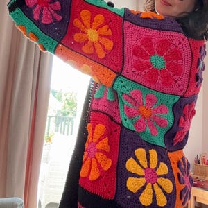 May include: A vibrant, hand-crocheted cardigan with large floral squares in pink, orange, purple, and green. The long-sleeved garment has a black border and a striped hem, showcasing a bohemian style. The cardigan is a statement piece.