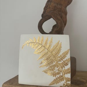 Könnte beinhalten: Eine weiße quadratische Keramikfliese mit einem goldenen Farnblatt-Design. Die Fliese liegt auf einem Holzregal.