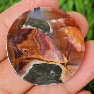 May include: A round, polished gemstone with a unique pattern of colors. The stone features a mix of brown, red, yellow, and green hues, creating a marbled effect. The gemstone is held in a hand, showcasing its size and detail.