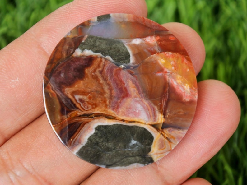 May include: A round, polished gemstone with a unique pattern of colors. The stone features a mix of brown, red, yellow, and green hues, creating a marbled effect. The gemstone is held in a hand, showcasing its size and detail.