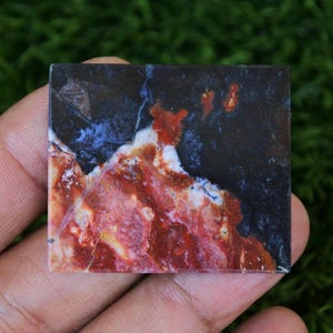May include: A rectangular gemstone with a striking pattern of red, white, blue, and black. The stone's surface displays a marbled effect, with the colours blending and swirling together. The gemstone is held in a hand, showcasing its natural beauty and unique colour variations.
