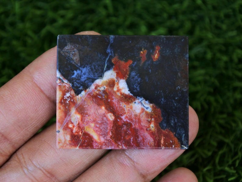 May include: A rectangular gemstone with a striking pattern of red, white, blue, and black. The stone's surface displays a marbled effect, with the colours blending and swirling together. The gemstone is held in a hand, showcasing its natural beauty and unique colour variations.
