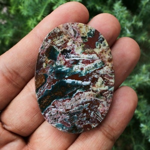 Puede incluir: Una piedra preciosa de forma ovalada con un patr&oacute;n de colores &uacute;nico. La piedra presenta una mezcla de tonos rojos, verdes, blancos y marrones, creando un efecto veteado. La superficie pulida de la piedra es lisa y refleja la luz.