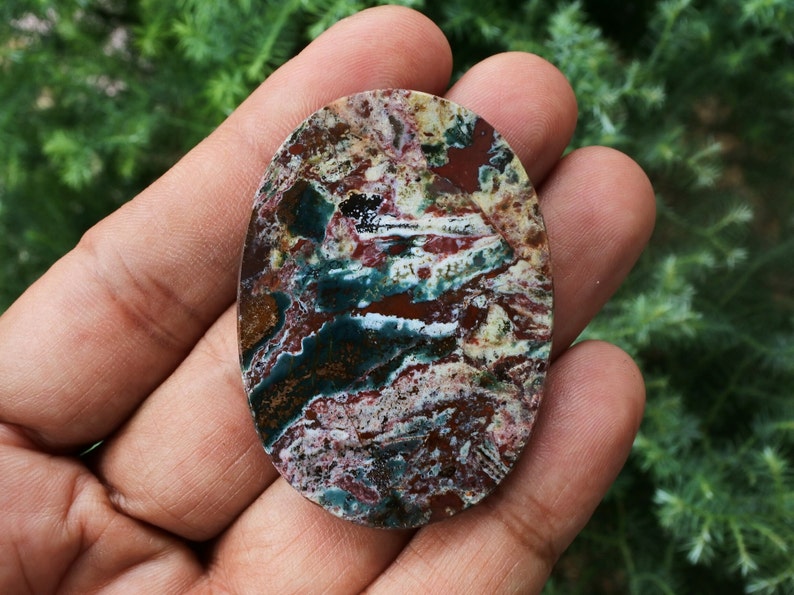 Puede incluir: Una piedra preciosa de forma ovalada con un patr&oacute;n de colores &uacute;nico. La piedra presenta una mezcla de tonos rojos, verdes, blancos y marrones, creando un efecto veteado. La superficie pulida de la piedra es lisa y refleja la luz.
