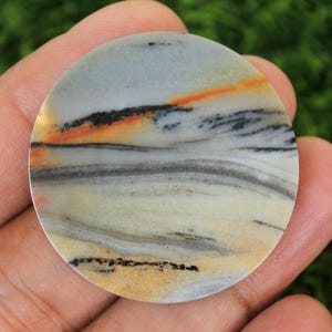 May include: A round, polished stone with a unique pattern of colors. The stone features a blend of light blue, cream, black, and orange hues, creating a landscape-like design. The stone is held in a hand, showcasing its natural beauty.