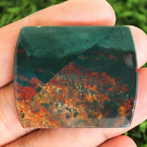 May include: A polished, rectangular gemstone with rounded corners. The stone features a dark green upper half and a lower half with red, orange, and brown patterns. The stone is held in a hand.