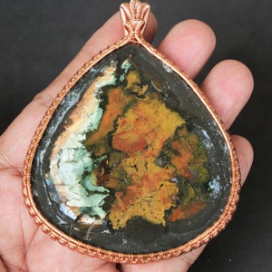 Peut inclure: Un pendentif en forme de goutte d'eau avec un fil de cuivre. La pierre a une base sombre avec des taches vertes, jaunes et oranges. Le pendentif est tenu dans une main sur un fond sombre. Le pendentif mesure environ 7.6 cm de long.