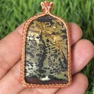 Może przedstawiać: Prostokątna zawieszka z miedzianą ramą drucianą. Zawieszka posiada kamień z mieszanką czarnych, złotych i zielonych wzorów. Drut jest misternie spleciony wokół kamienia, tworząc dekoracyjną obwódkę i pętlę na łańcuszek.