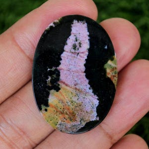 Puede incluir: Gema ovalada con un patrón llamativo. La piedra presenta una banda vertical central de color rosa y blanco, enmarcada por áreas negras. Toques de amarillo y verde realzan su apariencia única. Una piedra natural pulida.