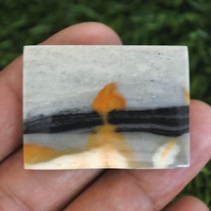 Op de afbeelding: Een rechthoekige gepolijste edelsteen cabochon met een glad oppervlak, voorzien van horizontale banden in lichtgrijs, een opvallende donkerzwarte streep en levendige oranje insluitingen die een duidelijk contrast vormen.