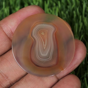 Può includere: Una pietra di agata rotonda e lucidata con bande concentriche di tonalità marrone, bianco e arancione. La pietra presenta un motivo centrale a forma di occhio ed è tenuta in mano su uno sfondo di erba verde. L'agata è una gemma naturale.