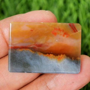 May include: A rectangular gemstone with a polished surface. The stone features a blend of colors, including orange, red, white, and gray. The colors are swirled together, creating a unique pattern. The gemstone is held in a hand.