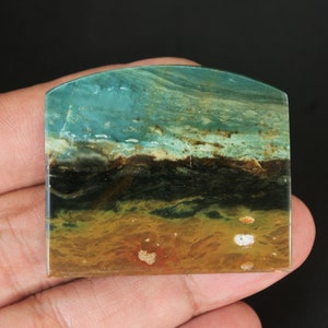 May include: A rectangular, polished gemstone with a curved top edge. The stone displays layers of teal, brown, black, and tan, reminiscent of a landscape. The stone is held in a hand against a black backdrop.