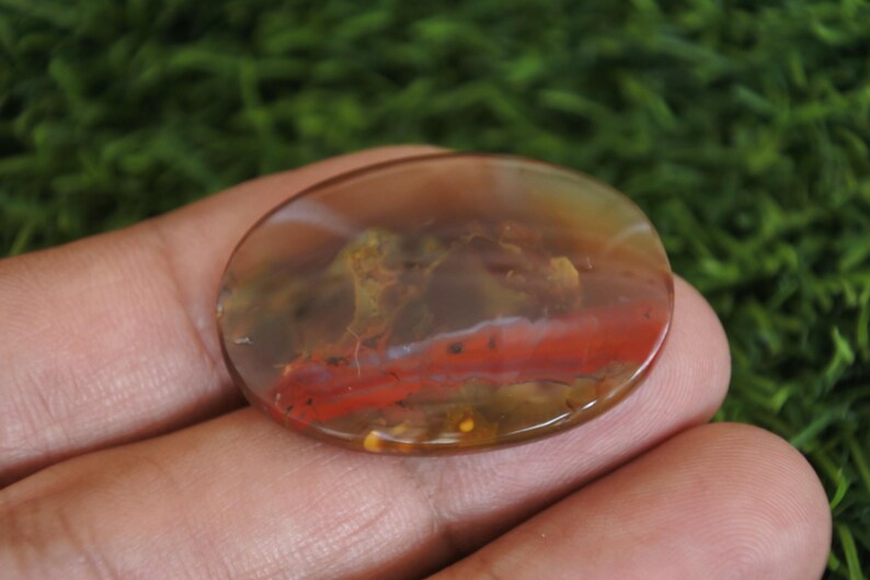 May include: An oval-shaped polished gemstone featuring a translucent brown and amber base with a distinct horizontal band of vibrant red color and internal moss-like inclusions throughout the smooth, rounded surface of the stone.