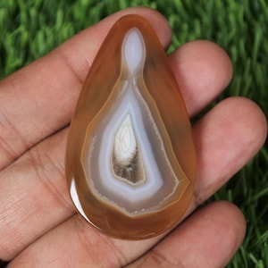 May include: A teardrop-shaped agate stone with concentric layers of brown, white, and tan hues. The polished gemstone is held in a hand, showcasing its natural patterns and colors. The stone's size is approximately 3 inches long.