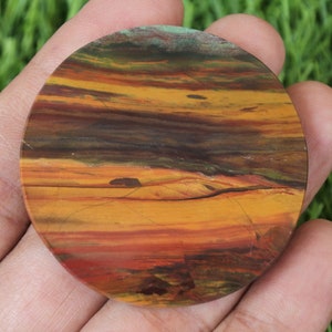 May include: A circular, polished stone displaying a natural, layered colour pattern. The stone has bands of orange, yellow, brown, and red, with touches of green. The round shape is held in a hand, highlighting the stone's unique, earthy hues.