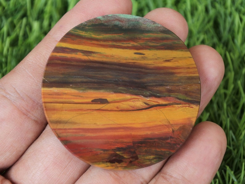 May include: A circular, polished stone displaying a natural, layered colour pattern. The stone has bands of orange, yellow, brown, and red, with touches of green. The round shape is held in a hand, highlighting the stone's unique, earthy hues.