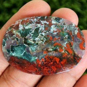 May include: An oval-shaped gemstone with a vibrant mix of colors. The stone features a base of deep red and green, with patches of white and purple. The polished surface highlights the natural patterns and textures of the stone.