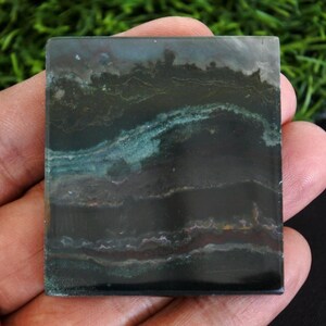 以下が含まれることがあります： 濃い緑色と黒色の正方形の磨かれた石。石は、ターコイズと茶色のヒントを持つ層状のパターンを持ち、自然でアースカラーの美しさを生み出しています。エッジはきれいで、石の滑らかな表面を示しています。