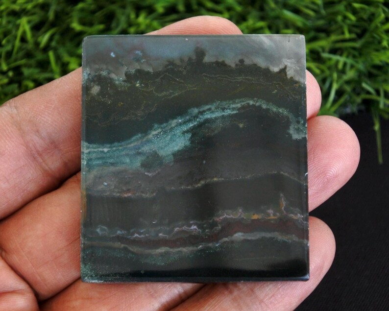 以下が含まれることがあります： 濃い緑色と黒色の正方形の磨かれた石。石は、ターコイズと茶色のヒントを持つ層状のパターンを持ち、自然でアースカラーの美しさを生み出しています。エッジはきれいで、石の滑らかな表面を示しています。