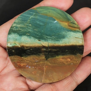 May include: A round, polished stone with a layered appearance. The top section is a mix of teal and green, followed by a dark brown band, and a bottom section of tan and brown. The stone is held in a hand.