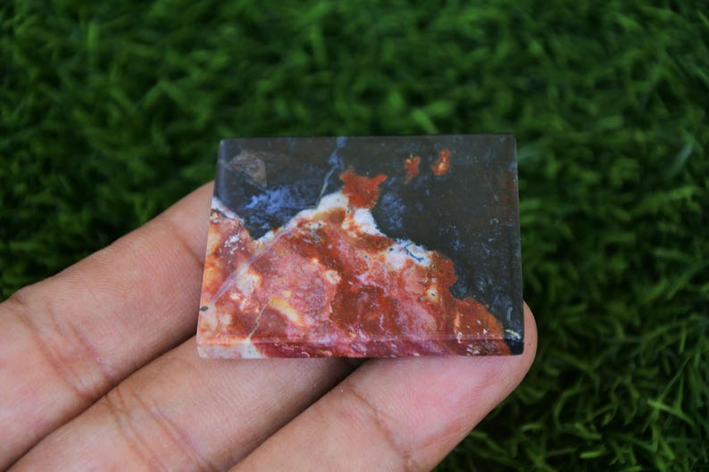May include: A rectangular stone with a unique pattern of red, white, blue, and black colours. The stone is held in a hand against a green background. The stone's surface has a marbled appearance.