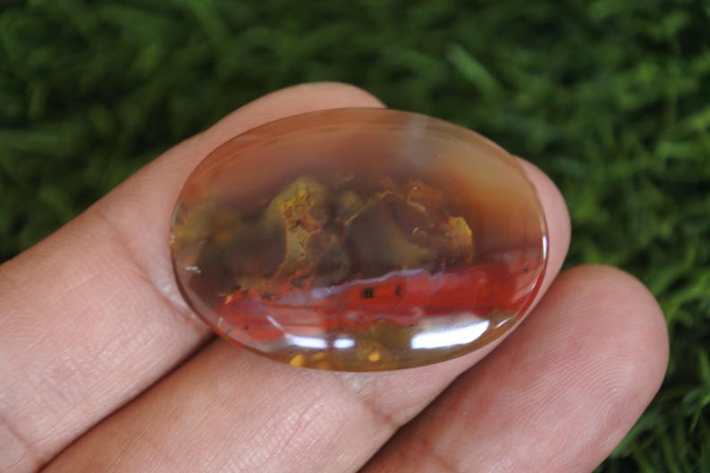 May include: An oval-shaped polished gemstone featuring a translucent amber and brown base with a distinct horizontal band of deep red and internal moss-like inclusions in shades of olive green and yellow throughout the smooth surface.