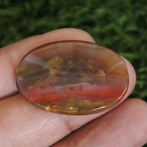 May include: An oval-shaped polished gemstone featuring a translucent brown base with a prominent horizontal band of vibrant red color and internal inclusions that appear as mossy green and yellow patterns throughout the smooth surface.