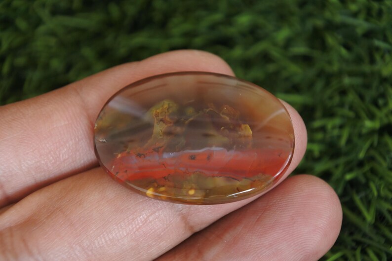 May include: An oval-shaped polished gemstone featuring a translucent brown base with a prominent horizontal band of vibrant red color and internal inclusions that appear as mossy green and yellow patterns throughout the smooth surface.