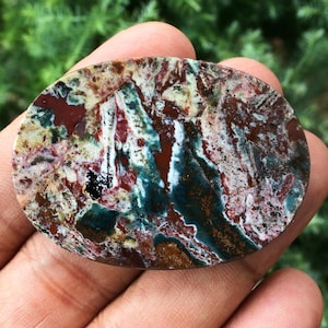 Può includere: Una gemma ovale con un motivo unico di tonalità rosse, verdi, bianche e marroni. La superficie lucida presenta un effetto marmorizzato, con aree di rosso intenso e verde acqua che contrastano con toni più chiari e cremosi. Una pietra naturale.