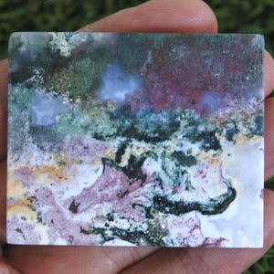 May include: A rectangular stone with a natural, abstract design. The stone features a blend of colors, including green, purple, white, and black, creating a landscape-like pattern. The stone is held in a hand, showcasing its size and texture.