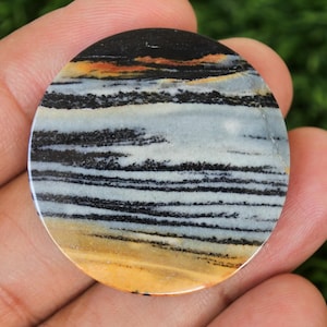 May include: A circular, polished stone with a layered design. The top section is black, transitioning to orange and yellow, then light blue and white with black stripes. The bottom is a light yellow colour.