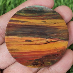 Peut inclure: Une pierre ronde et polie avec un motif de couleurs en couches. La pierre présente des bandes orange, jaune, marron et verte, ressemblant à un coucher de soleil. La pierre est tenue dans une main.