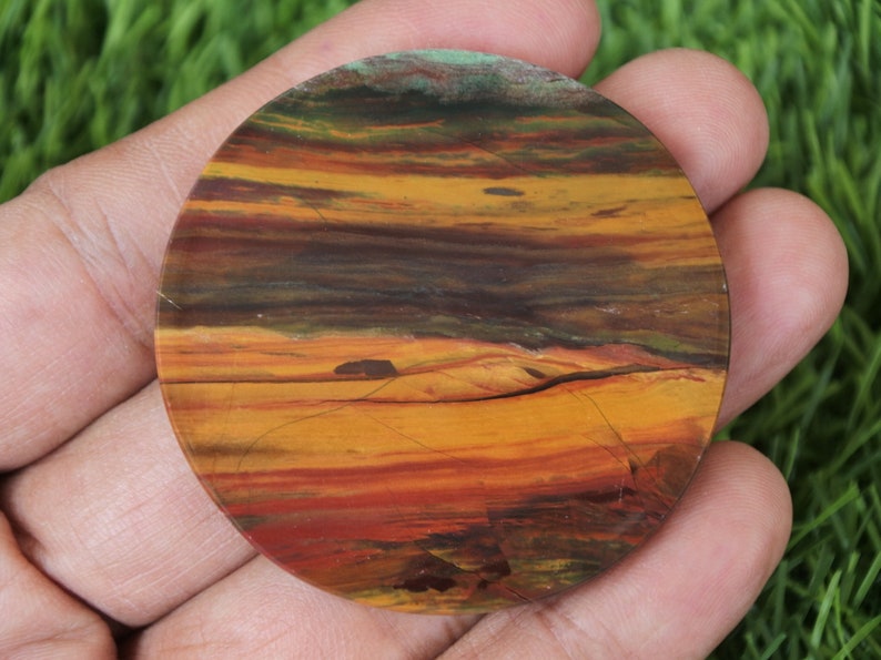 May include: A circular, polished stone displaying a layered pattern of colours. The stone has bands of orange, yellow, brown, and green, reminiscent of a sunset. The stone is held in a hand.