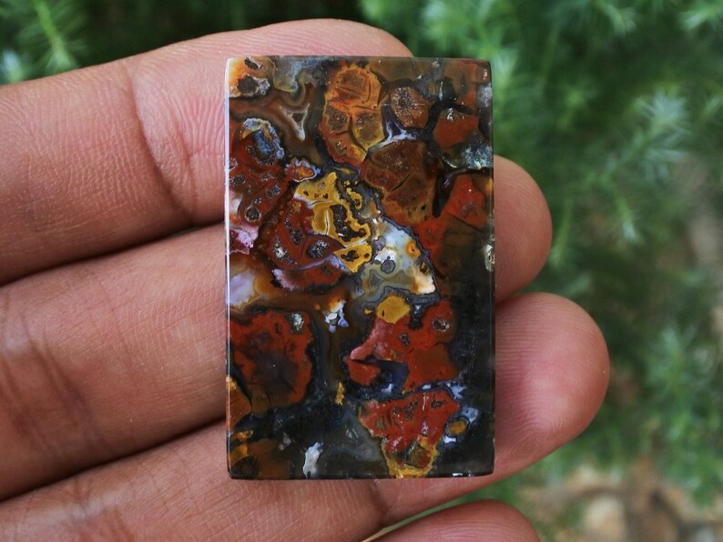 Op de afbeelding: Een rechthoekige, gepolijste agaat cabochon met een kleurrijk, abstract patroon van rood, oranje, geel, bruin en zwart. De agaat heeft een gladde, glanzende afwerking.