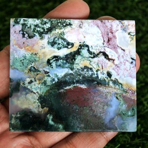May include: A square, polished stone with a unique, natural pattern. The stone features a blend of colors, including green, white, pink, and brown, creating a marbled effect. The stone is held in a hand, showcasing its intricate details and textures.