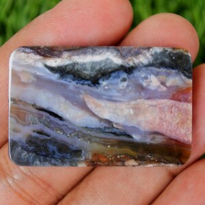 Pode incluir: Uma pedra preciosa retangular com uma superf&iacute;cie polida. A pedra apresenta uma mistura de cores, incluindo branco, preto, azul, roxo e castanho. O padr&atilde;o assemelha-se a uma paisagem com texturas em camadas e forma&ccedil;&otilde;es naturais. A pedra preciosa &eacute; segurada em uma m&atilde;o.